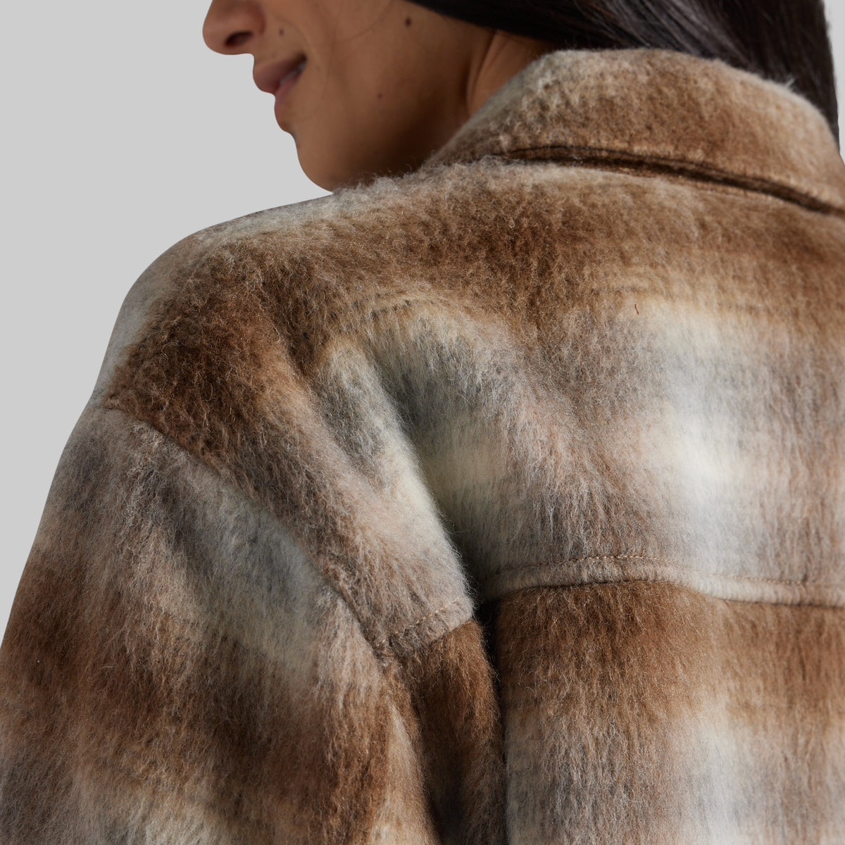 Close-up of a person wearing a soft, fuzzy faux-fur or shearling-style jacket, showing the back and shoulder area with a brown, beige, and gray marled pattern against a plain light gray background.