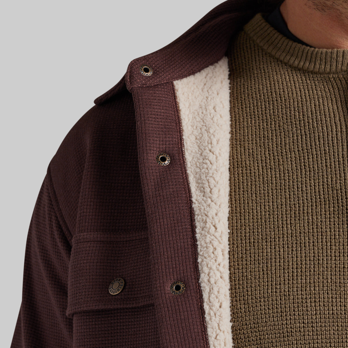 Close-up of a person wearing a dark brown waffle-knit jacket with a cream sherpa lining over an olive green ribbed sweater, against a light gray background.