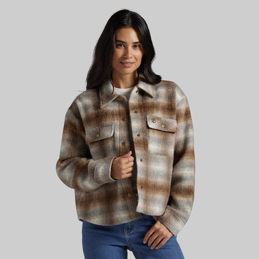 A woman modeling a cropped, long-sleeve plaid button-up jacket in brown, beige, and gray tones, paired with blue jeans, against a solid gray studio background.