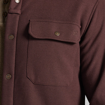 Close-up of a waffle-knit button-front shirt jacket in a muted burgundy-brown color, featuring a chest pocket with a flap and metal snap, plus a small embroidered logo.