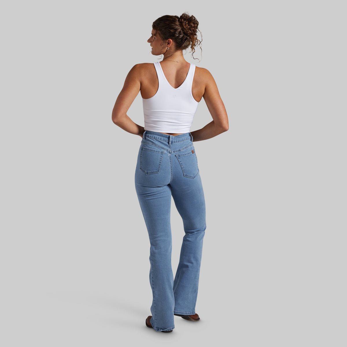 A woman is shown from the back wearing a white tank top and light blue high-waisted bootcut jeans, standing against a solid gray studio background.