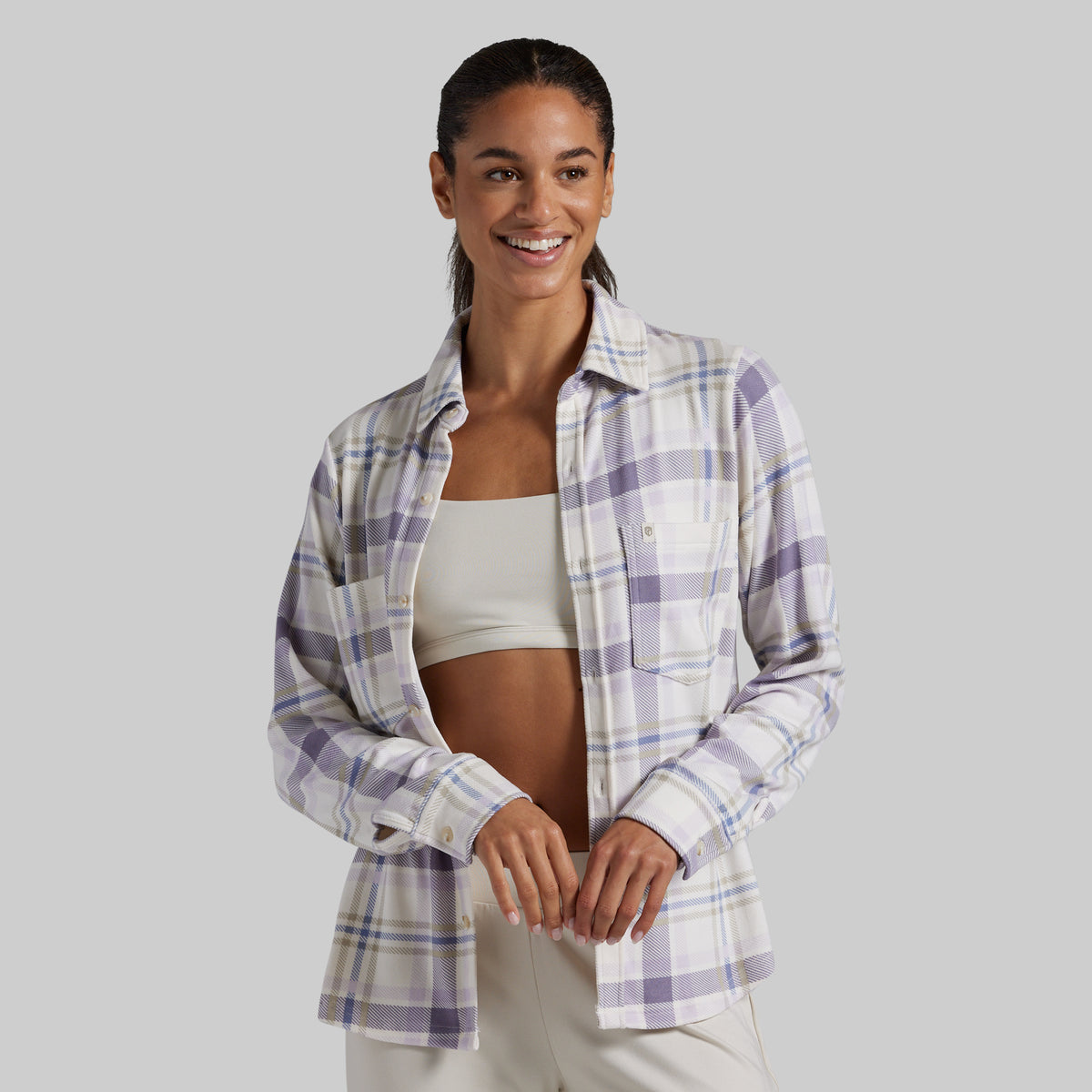 A woman modeling a long-sleeve plaid button-up shirt in lavender, white, and light blue over a beige sports bra, set against a solid gray studio background.