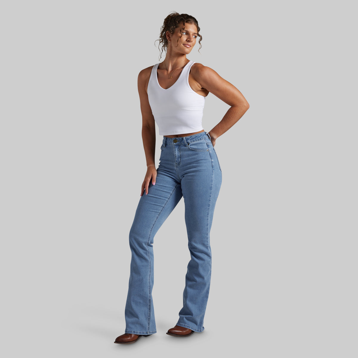A woman modeling light-wash blue bootcut jeans with a white ribbed tank top, standing against a solid gray studio background.