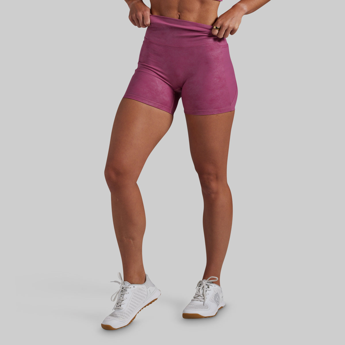 A model wearing high-waisted athletic shorts in a dusty rose or mauve color, paired with white sneakers, shown from the waist down against a solid gray background.