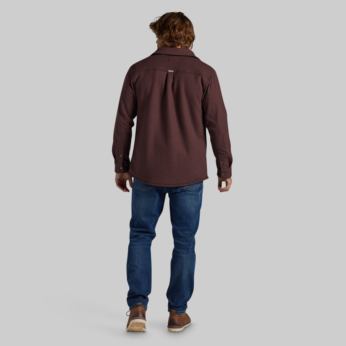 A man viewed from behind wearing a dark brown long-sleeve button-up shirt, blue jeans, and brown shoes, standing against a solid gray background.