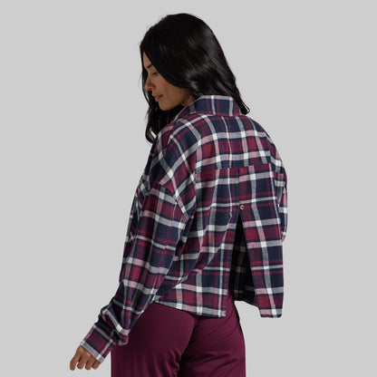 A woman wearing a cropped, long-sleeve plaid flannel shirt in burgundy, navy, and white, paired with burgundy pants, shown from a side-back angle.