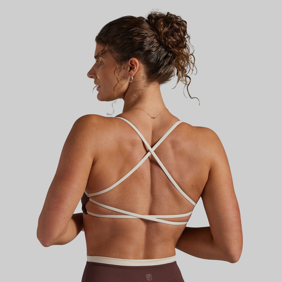 A woman shown from the back wearing a sports bra with thin crisscrossing straps in a light beige color, paired with a brown sports bottom, against a solid gray background.