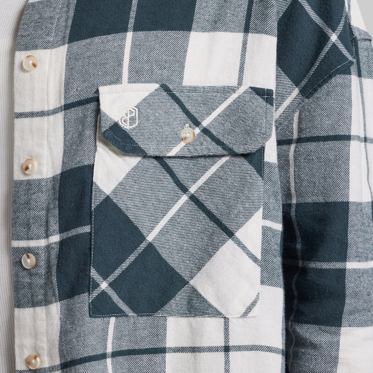 Close-up of a plaid button-front shirt with a chest pocket, featuring dark teal, white, and light gray diagonal checks. A small embroidered shield logo is visible on the pocket flap.