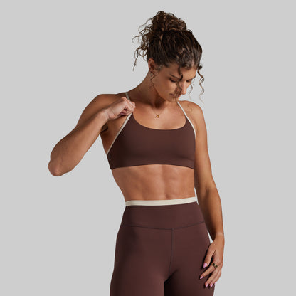 A woman modeling a dark brown activewear set consisting of a sports bra and high-waisted leggings, featuring a contrasting light beige waistband and underband, set against a solid gray background.