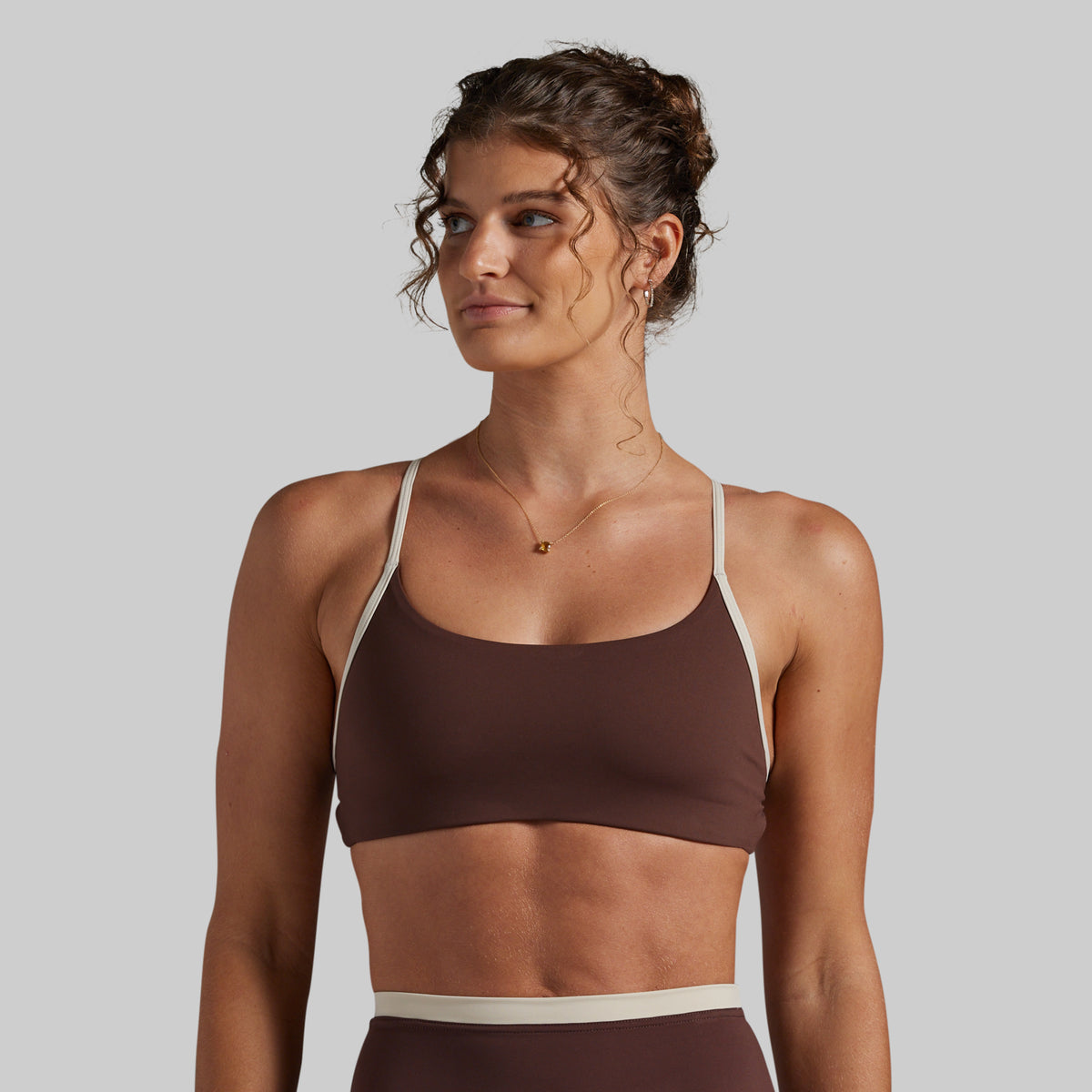 A woman wearing a brown sports bra with contrasting beige straps and a matching beige waistband, shown against a solid light gray background.