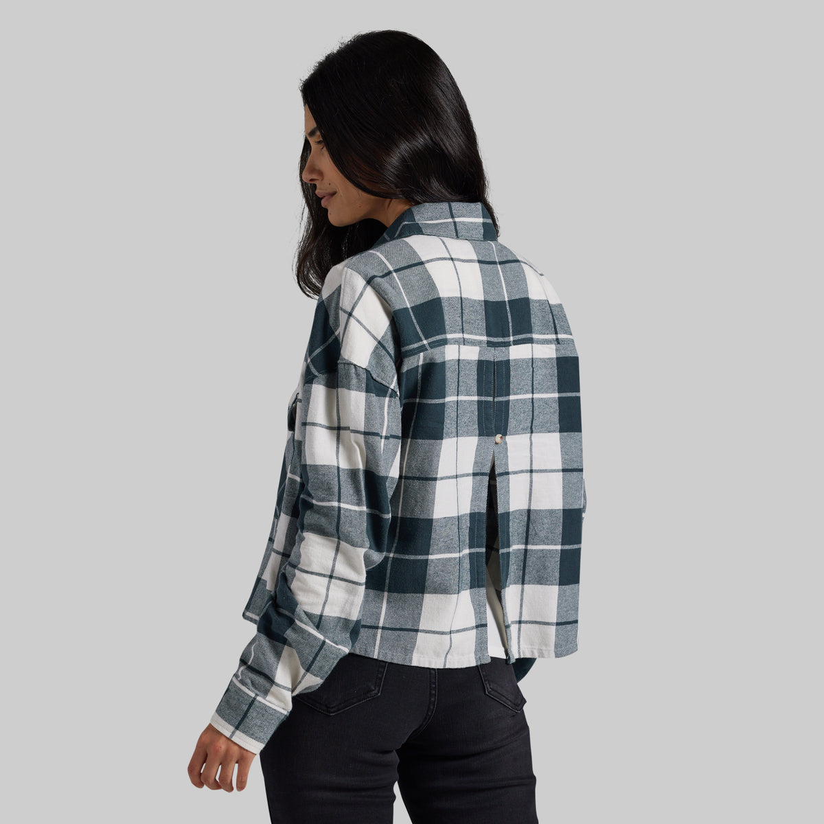 A woman shown from the back wearing a long-sleeve plaid button-up shirt in dark teal and white, paired with black jeans, against a solid light gray background.