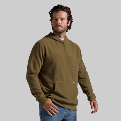 A man wearing an olive green pullover hoodie with a front kangaroo pocket, paired with blue jeans, standing against a solid gray background.