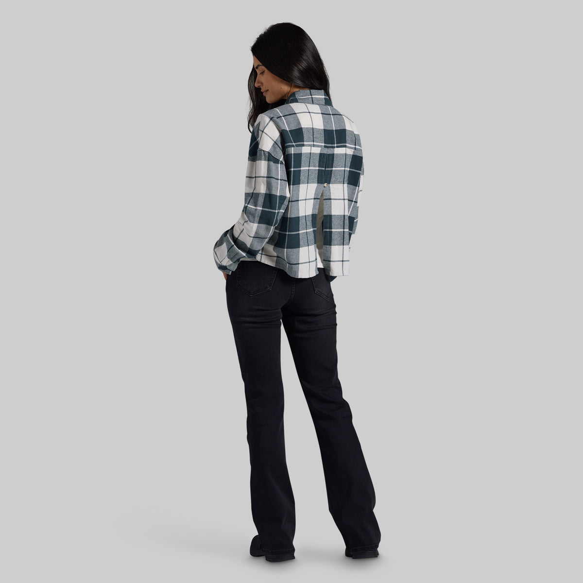 A woman shown from the back wearing a dark green and white plaid flannel shirt and black jeans, standing against a solid gray studio background.
