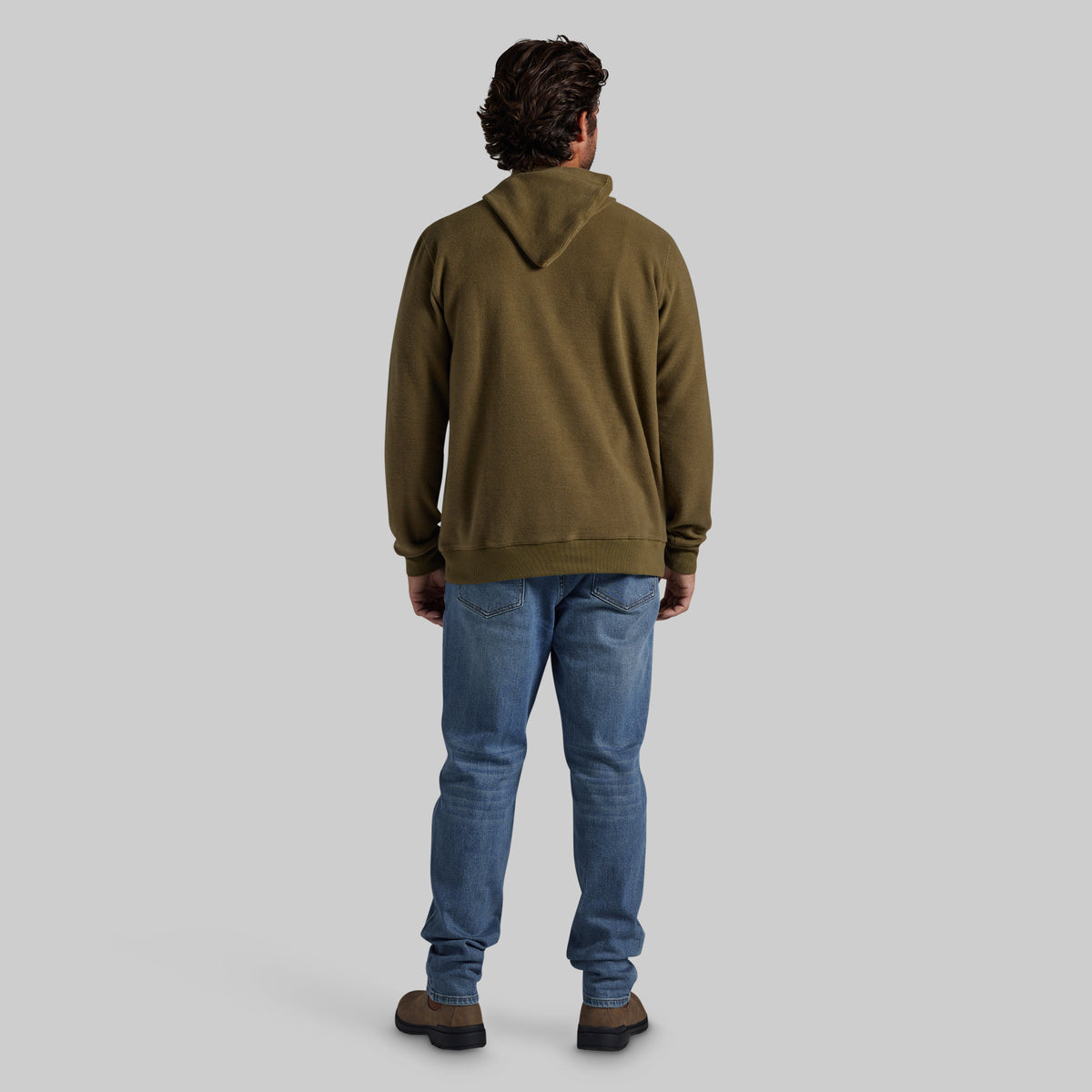 A man viewed from behind wearing an olive green pullover hoodie, blue jeans, and brown shoes, standing against a solid light gray background.