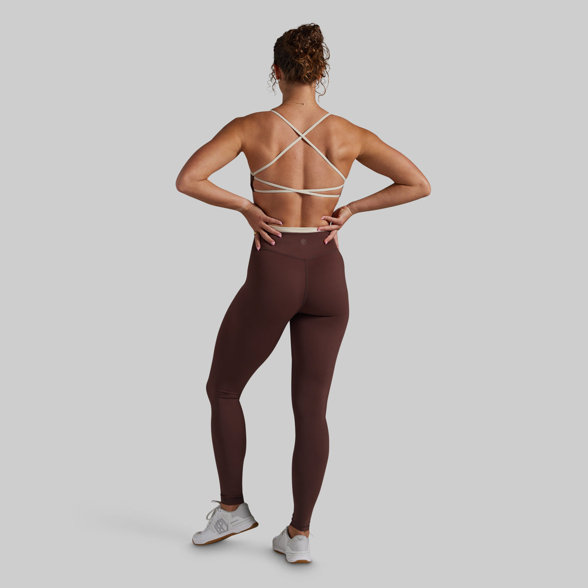 A woman shown from the back wearing a dark brown activewear set consisting of a strappy sports bra and high-waisted leggings, paired with white sneakers, against a solid gray background.