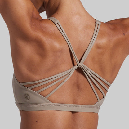 Rear view of a person wearing a beige sports bra with multiple thin crisscross back straps, shown against a light gray studio background.