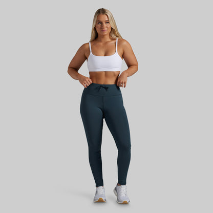 A woman modeling high-waisted athletic leggings in a dark teal color, paired with a white sports bra and white sneakers, against a solid light gray background.
