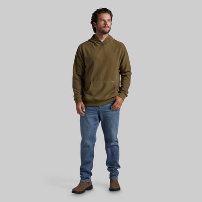 A man wearing an olive green pullover hoodie, blue jeans, and brown boots, standing against a solid gray studio background.