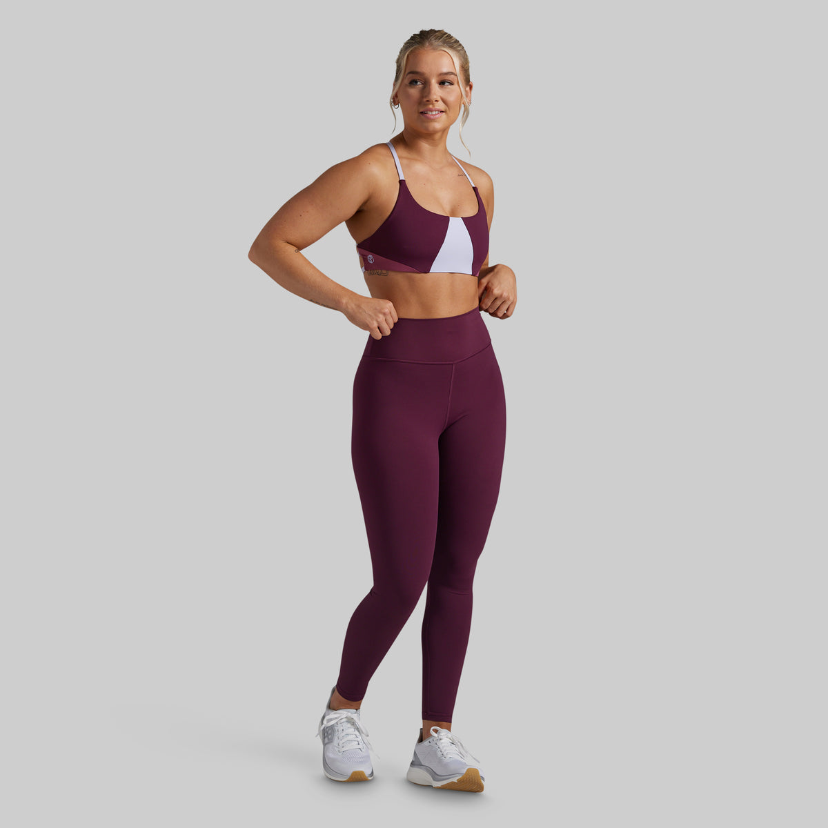 A woman modeling a matching athletic set consisting of a color-blocked sports bra and high-waisted leggings in maroon with a white accent panel, paired with white sneakers, against a solid gray background.