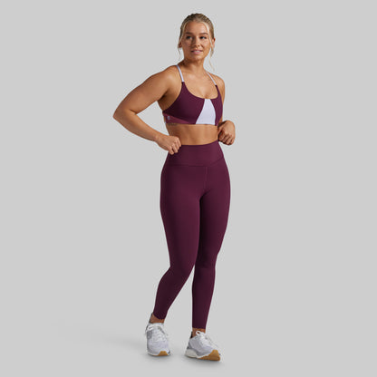 A woman modeling a matching athletic set consisting of a color-blocked sports bra and high-waisted leggings in maroon with a white accent panel, paired with white sneakers, against a solid gray background.