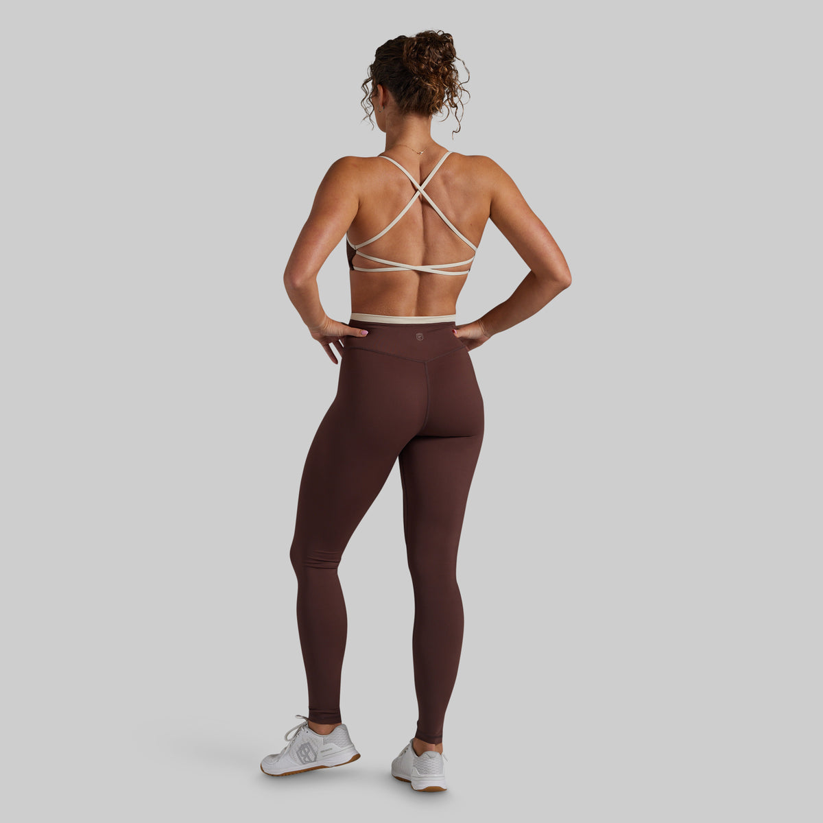 A woman shown from the back wearing a brown athletic leggings set with a matching sports bra featuring cream-colored crisscross straps, posed against a solid light gray background.