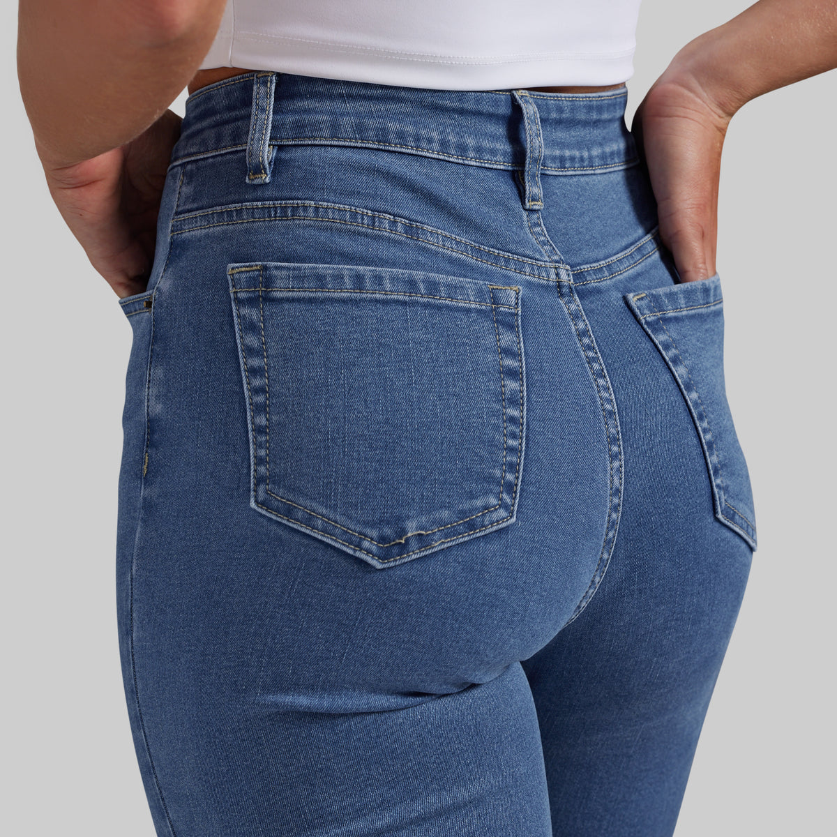 Close-up back view of a person wearing blue denim jeans with a white cropped top, showing the back pockets and waistband details against a light gray studio background.