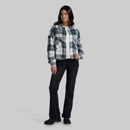 A woman modeling a long-sleeve plaid button-up shirt in black, white, and gray, worn over a white t-shirt and paired with black jeans and black shoes, against a solid gray studio background.