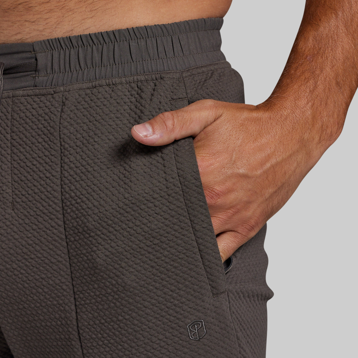 Close-up of a person wearing textured dark gray athletic joggers, showing the side pocket with a hand inserted and an elastic waistband.