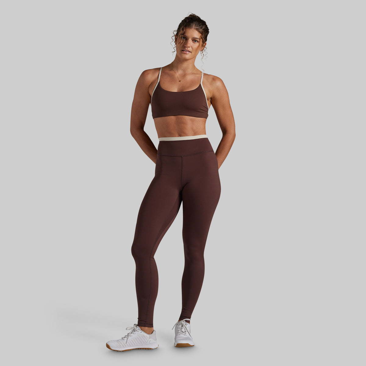 A woman modeling a matching athletic set consisting of a dark brown sports bra and high-waisted leggings with a contrasting light beige waistband, paired with white sneakers, against a solid light gray background.