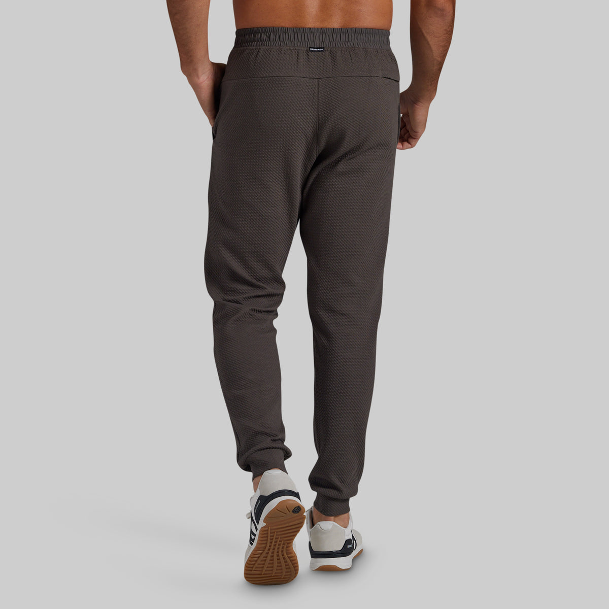 Rear view of a person wearing dark greay textured jogger pants with elastic cuffs, paired with white and navy sneakers, against a light gray studio background.