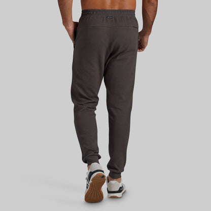 Rear view of a person wearing dark greay textured jogger pants with elastic cuffs, paired with white and navy sneakers, against a light gray studio background.