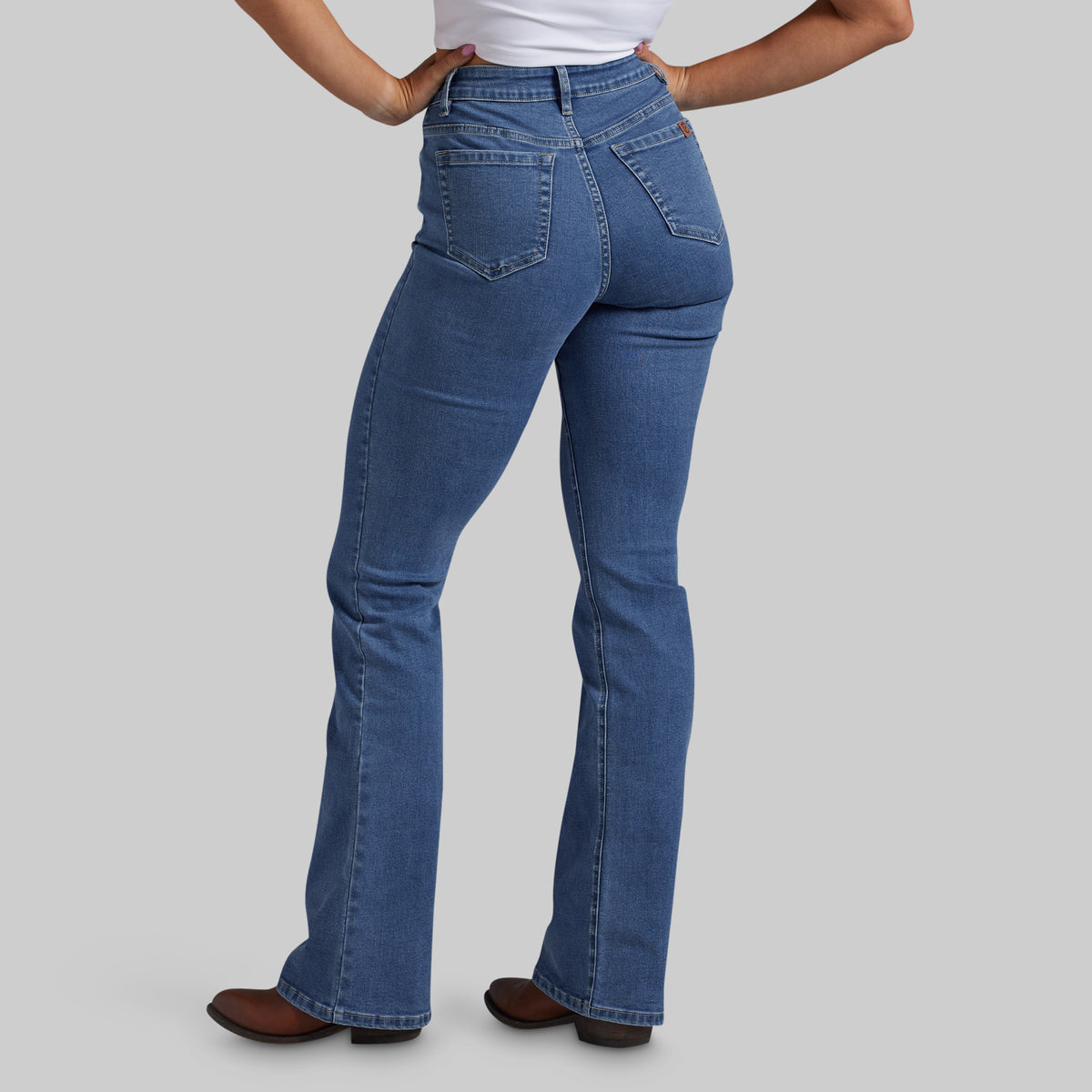 A person shown from the back wearing blue denim jeans with a straight-to-flare leg, paired with a white top and brown shoes, against a solid gray studio background.