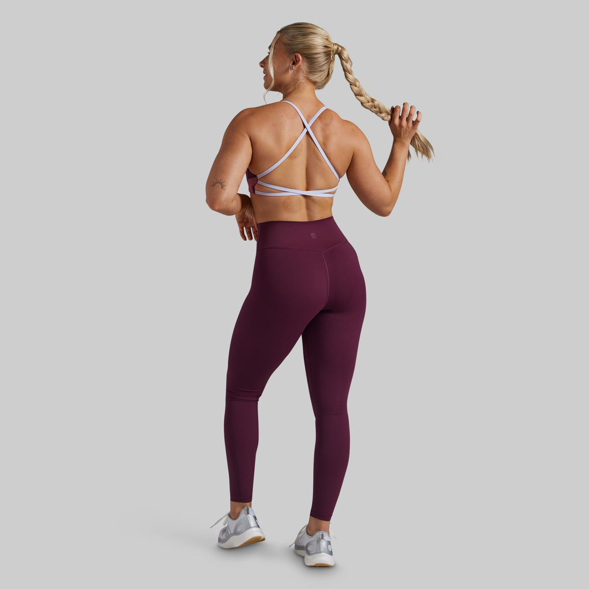 A woman shown from the back wearing a high-waisted, form-fitting burgundy athletic leggings and a matching strappy sports bra, standing against a solid light gray background.