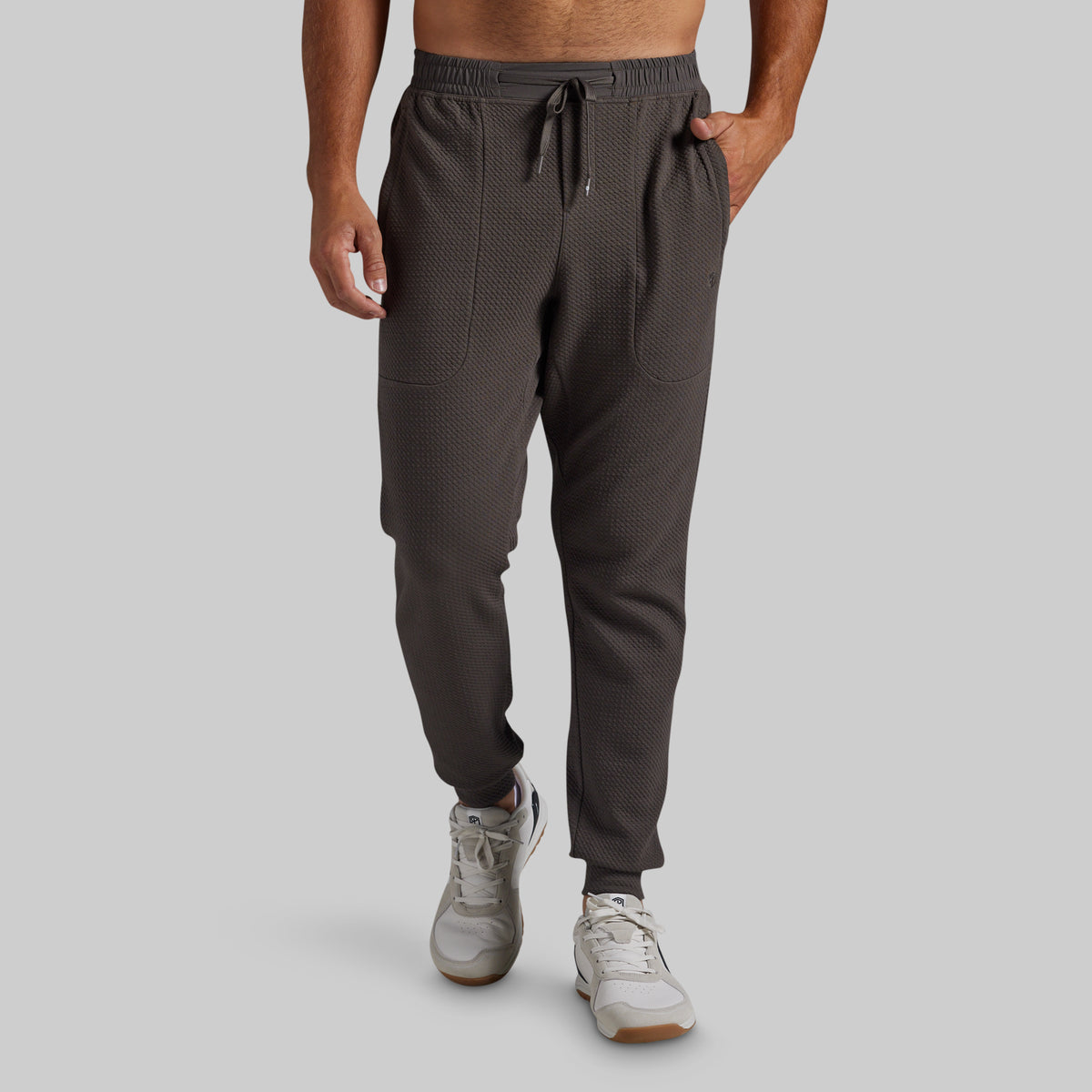 A man wearing dark gray textured jogger pants with a drawstring waist and side pockets, paired with white sneakers, shown from the waist down against a solid gray background.
