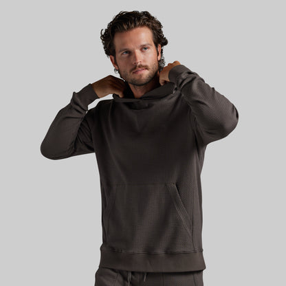 A man wearing a dark grey textured pullover hoodie with a high neck and kangaroo pocket, posing with his hands behind his neck against a solid gray background.