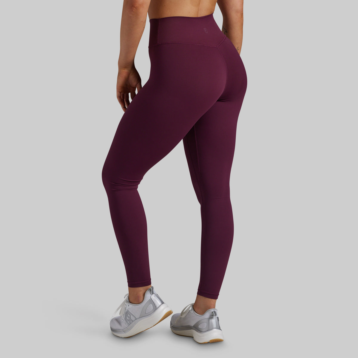 A model from the back wearing high-waisted, form-fitting leggings in a deep plum purple color, paired with silver sneakers, against a solid light gray background.