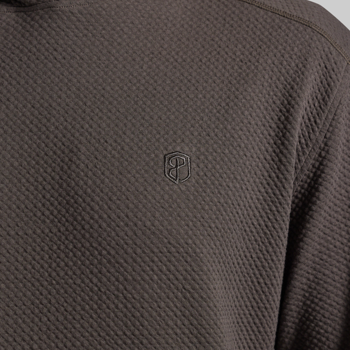 Close-up of a dark grey textured hoodie with a small tonal embroidered shield logo on the chest.