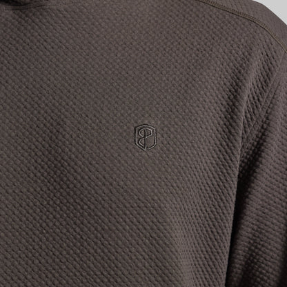 Close-up of a dark grey textured hoodie with a small tonal embroidered shield logo on the chest.