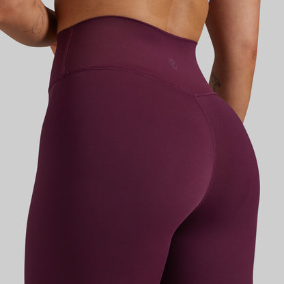 Close-up rear view of a person wearing high-waisted leggings in a deep plum-purple color against a light gray studio background.