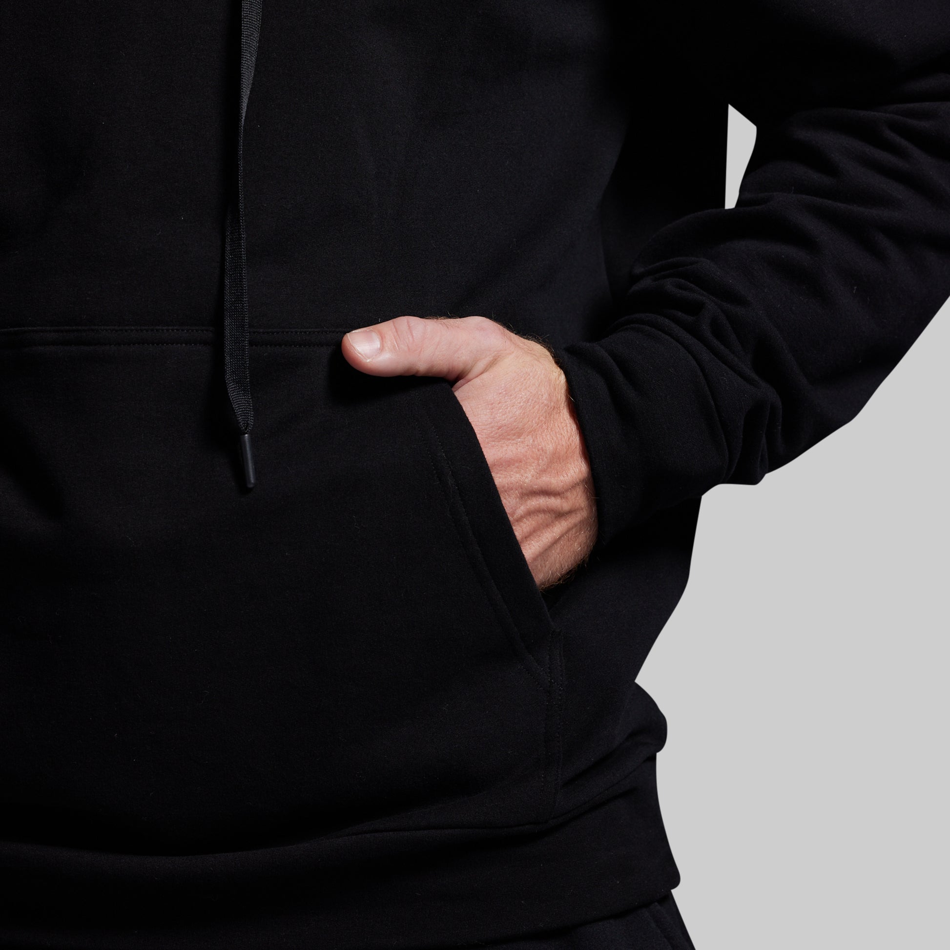 Person wearing a black hoodie with a hand in the pocket on a light gray background