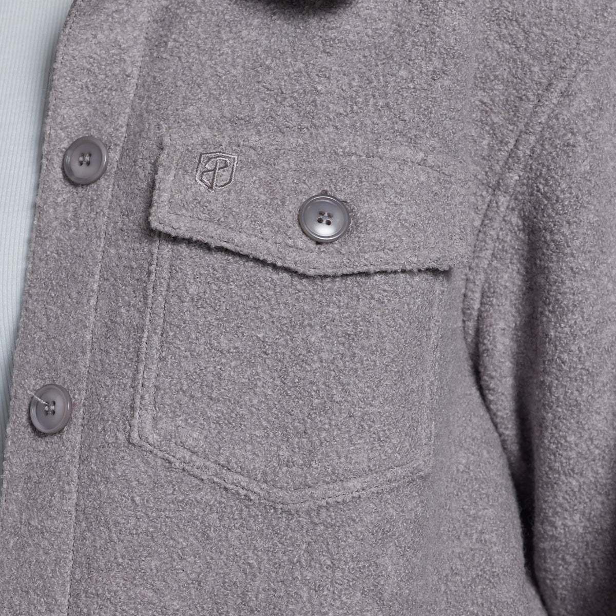 Close-up of a textured gray button-up jacket with a chest pocket, showing a small shield logo on the pocket flap.