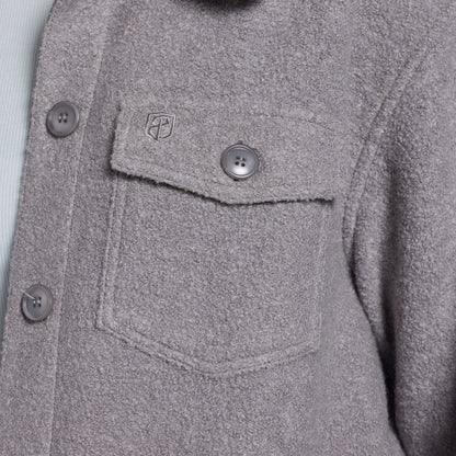 Close-up of a textured gray button-up jacket with a chest pocket, showing a small shield logo on the pocket flap.