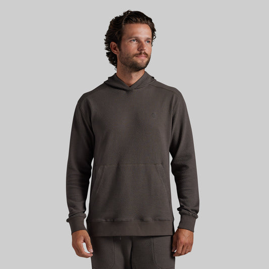 A man wearing a dark brown/grey textured waffle-knit hoodie and matching sweatpants, standing against a solid light gray background.