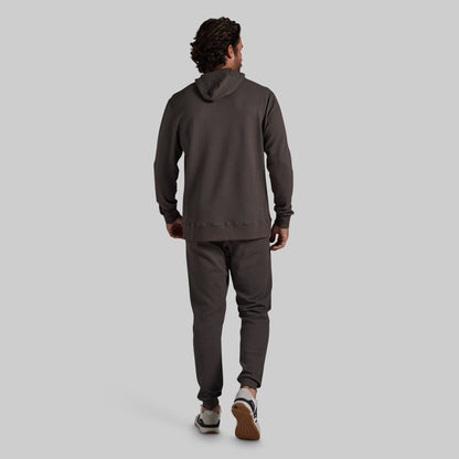 A man is shown from behind wearing a dark grey hooded sweatshirt and matching jogger pants, walking against a solid light gray background.