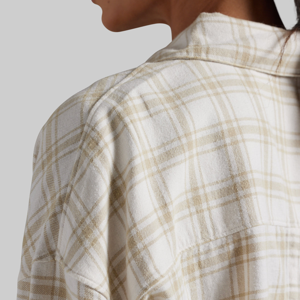 Close-up of a person wearing a cream and beige plaid flannel shirt, showing the collar, shoulder, and fabric texture against a light gray background.