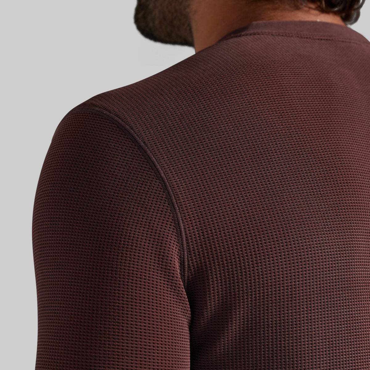 Close-up back view of a person wearing a textured, waffle-knit long-sleeve shirt in a deep burgundy brown color, set against a solid light gray background.
