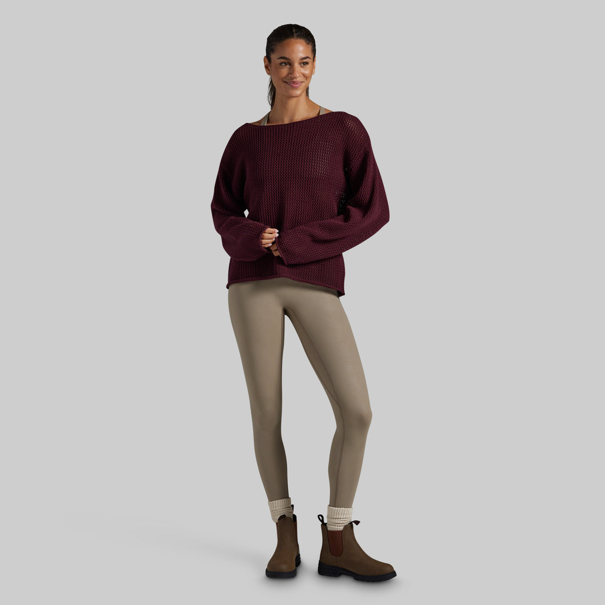 A woman modeling a dark burgundy ribbed knit sweater with a wide boat neckline, paired with beige leggings and brown Chelsea boots, against a solid light gray background.