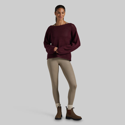 A woman modeling a dark burgundy ribbed knit sweater with a wide boat neckline, paired with beige leggings and brown Chelsea boots, against a solid light gray background.