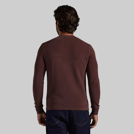 A man viewed from behind wearing a dark brown long-sleeve crewneck shirt and dark blue jeans, set against a solid light gray background.