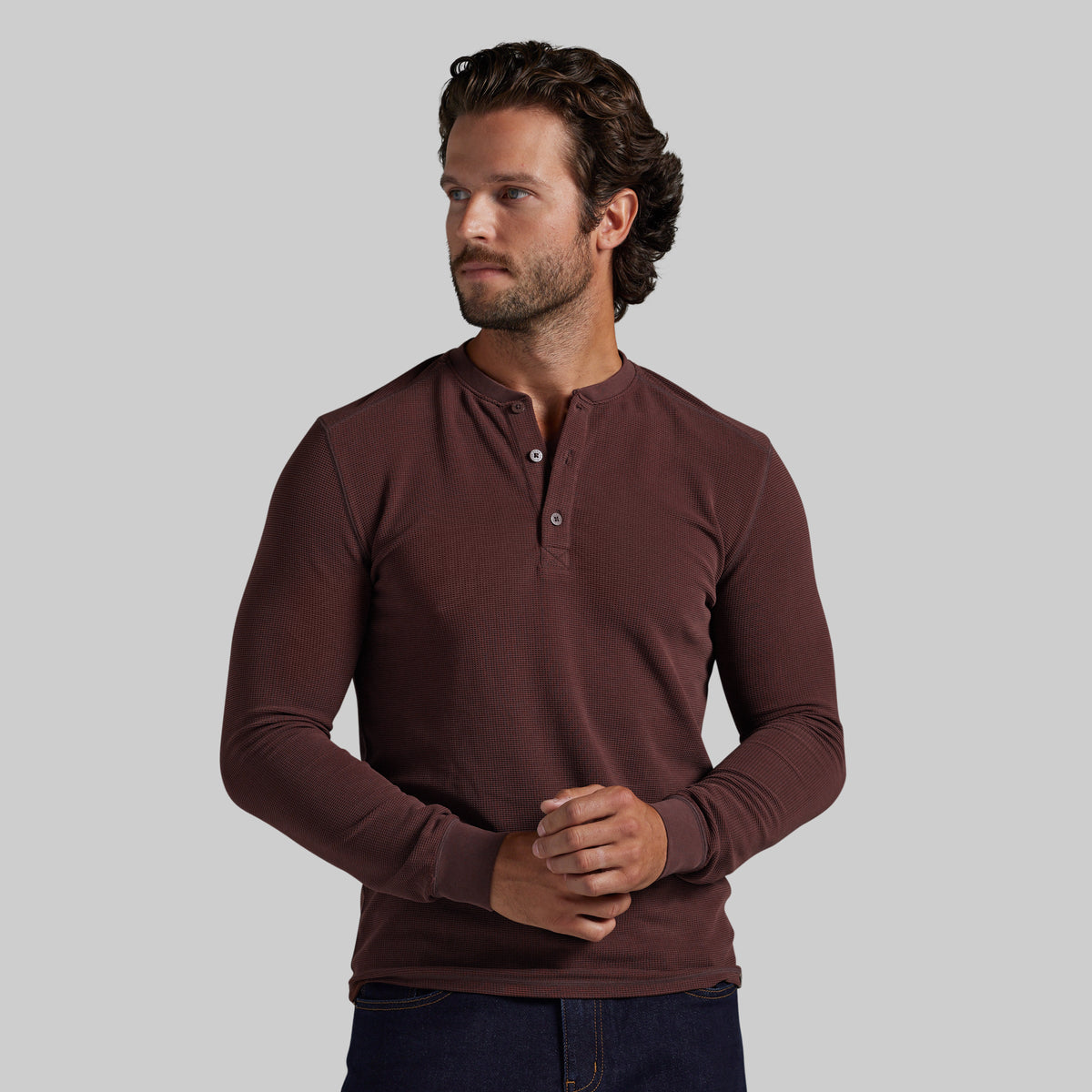 A man wearing a long-sleeve henley shirt in a deep burgundy brown color, paired with dark blue jeans, standing against a solid light gray background.